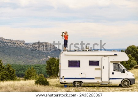 Woman enjoying trip with motor home, standing on roof of camper vehicle, holds camera, taking travel picture from mountain nature. Happiness, vacation and traveling.