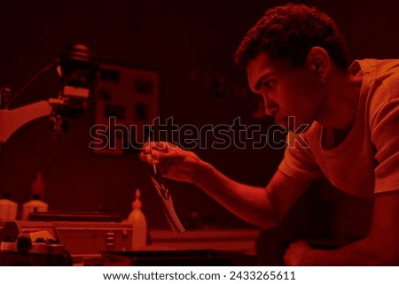 african american photographer holding developed photo paper with tweezers in red toned darkroom