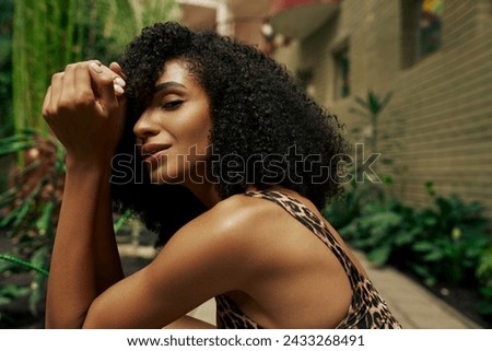 beautiful african american curly woman in chic attire smiling and posing around green plants Royalty-Free Stock Photo #2433268491