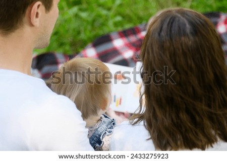 Parents reading a book to their child, sitting on a blanket during a picnic in the park, watching educational pictures, back view, close-up. Weekend in nature. Concept of happy family, learning