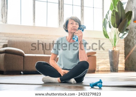 Tired exhausted caucasian senior woman relaxing after training on fitness mat in lotus position. Grandmother drinking water after home workout