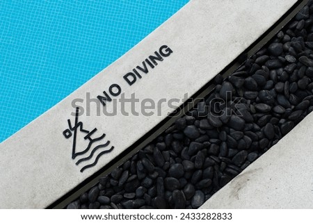 No Diving text warning sign and rules icon at a pool edge with a pictogram beside a blue tiled swimming pool and decorative black stones exterior design