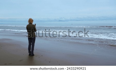 Woman on shore takes pictures of sea on phone. Clip. Woman takes pictures of sea on cloudy day. Woman takes pictures of sea with waves