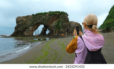 Woman on shore takes pictures of sea on phone. Clip. Woman photographs seashore with rocky arch. Group of tourists takes pictures of rocky arch on seashore