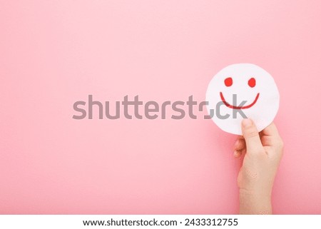 Baby girl hand holding drawn red happy smiling face on white paper on light pink table background. Pastel color. Closeup. Children mood concept. Empty place for text. Top view.