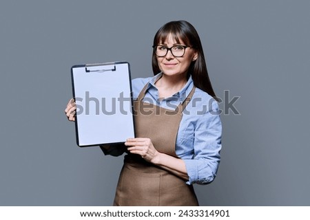 Smiling woman worker, owner in apron showing blank sheet of paper on clipboard Royalty-Free Stock Photo #2433314901