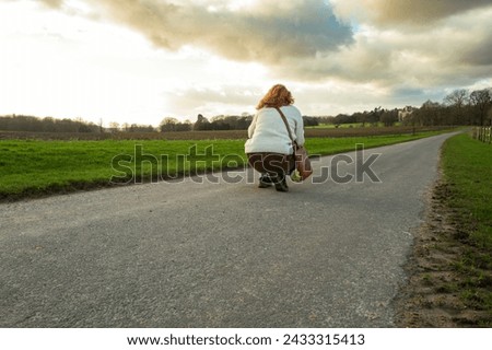 Auburn haired woman seen crouching in the middle of an empty road with the aim of taking landscape picture in early spring in the UK.