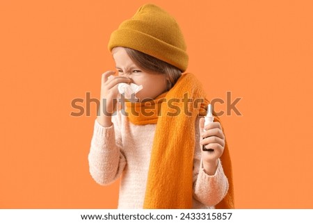 Sick little girl in hat with tissue and nasal drops sneezing on orange background