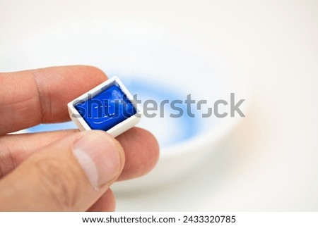 Landscape of A hand shows a mini blue watercolor cube with some more cubes of the same color palette and a container with water in the background out of focus.