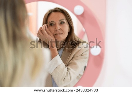 Elegant senior woman applying blush with a makeup brush, focused on her reflection. She wears a stylish striped shirt, chic short blonde hairstyle, exuding timeless beauty and confidence.