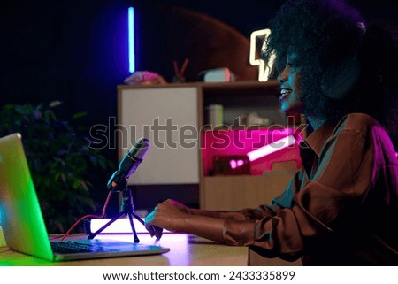 Young smiling African woman sitting in home office in headphones, with microphone and laptop, recording online podcast. Social media business. Concept of journalism, modern technology, mass media