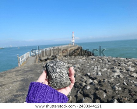 A woman's hand holding a geocache that looks like a black stone which was hidden between rocks on a beach on a sunny day in Netherlands Royalty-Free Stock Photo #2433344121