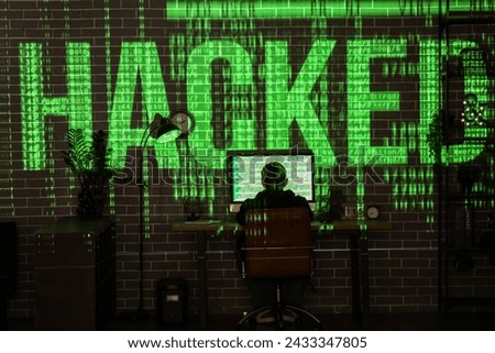 Hacker using computer at table and projection of word HACKED in dark room
