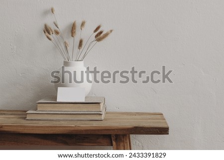 Blank business card mockup on old books. Modern round ceramic vase with dry bunny tails, Lagurus ovatus grass on wooden table, bench.. White wall background. Scandinavian interior. Home office.