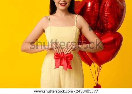 Beautiful young happy woman with gift box and heart-shaped balloons on yellow background. Valentine's Day celebration