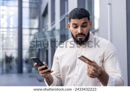 Close-up photo of a young Indian man in a shirt standing outside, holding a mobile phone and looking confusedly at a credit card.
