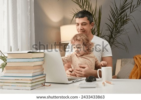 Smiling cheerful Caucasian man wearing casual clothing sitting at table in front of laptop with his infant baby daughter watching cartoons online together in home interior