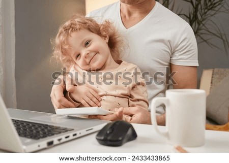 Smiling cute wavy haired toddler girl sitting at table in front of laptop with his father watching cartoons with dad while man having break of freelance work posing in home interior