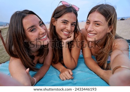 Three cheerful friends lying on a beach towel using phone to take a selfie looking happily at the camera. Female students having fun together on summer vacation. Girls trip.