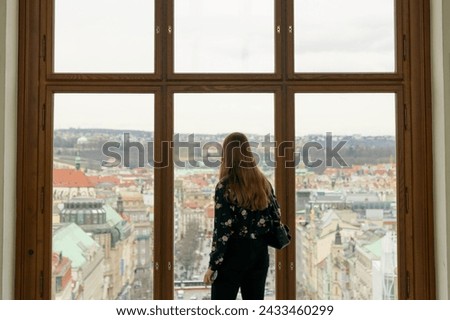 Woman overlooking Prague City - Large picture window Beautiful contrast of wood dark clothes red hair and gray sky