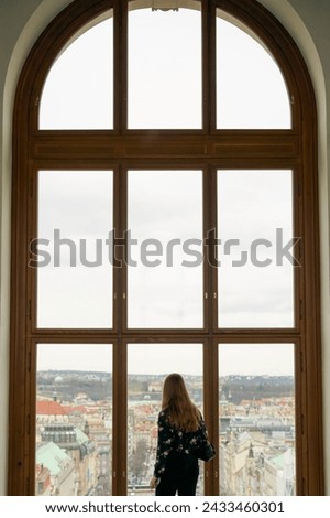Woman overlooking Prague City - Large picture window Beautiful contrast of wood dark clothes red hair and gray sky