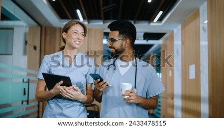 Portrait of skilled happy doctors walking in modern hospital hallways smiling and talking about their jobs. Caucasian doctor and young cheerful male Indian nurse having coffee.