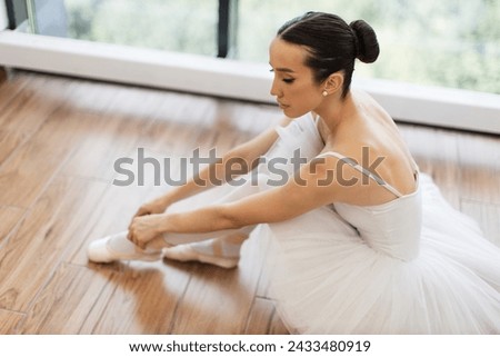 Beautiful young female ballet dancer relaxing after rehearsal. Side view portrait of talented ballerina finished her wonderful performance and resting while sitting at wooden floor, copy space. Royalty-Free Stock Photo #2433480919