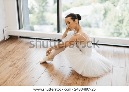 Side view portrait of talented ballerina finished her wonderful performance and resting while sitting at wooden floor, copy space. Beautiful young female ballet dancer relaxing after rehearsal. Royalty-Free Stock Photo #2433480925