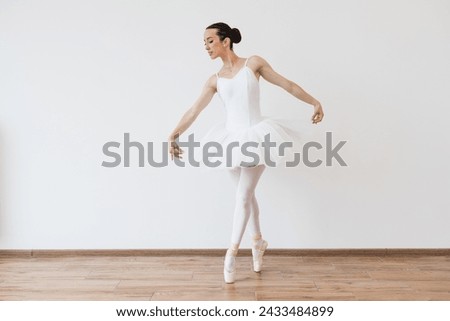 Beautiful young female dancer posing on studio background. Caucasian ballerina in white bodysuit and tutu poses in motion showing ballet elements while standing on pointe shoes.