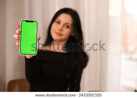 Smiling girl show smartphone with green chromakey screen, recommendation app promotion or mobile technology use in cafe. Online services, connectivity, ad and offer, sale. Focus on mobile phone.