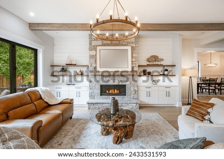 Living room interior with exposed wooden beams on the ceiling carmel colored leather sofa round wagon wheel chandelier fluffy white throw blanket hardwood floor view to kitchen and stone fireplace
