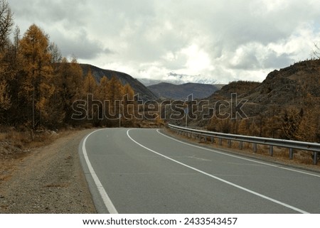 A two-lane asphalt road turns in an autumn valley towards the mountains on an overcast cloudy day. Chuisky tract, Altai, Siberia, Russia.