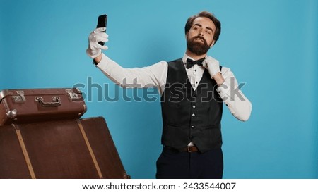 In studio, doorman in suit takes photos while using smartphone app for selfies and acting silly against blue background. Elegant professional hotel concierge doing pictures with his phone.
