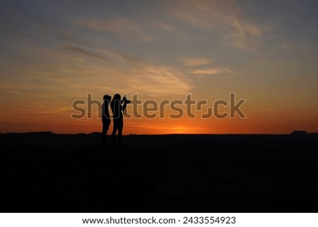Silhouetted couple taking photos of a serene sunset in a vast desert landscape, embodying a moment of adventure and tranquility.