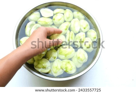 woman hand holding a Cut in half and peel green raw organic Mangoes (Mangifera indica L. Var. ) fruit in a bowl with water isolate on a backdrop. Prepare to do fruit compote , pickling mangoes, 