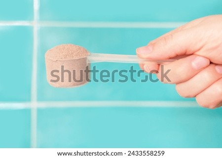 Female hand holding scoop with a brown protein on mint tile background, side view.