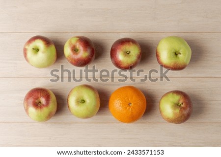 Top view of six apples in two rows with one distinct orange in the middle on a wooden surface, symbolizing uniqueness and individuality