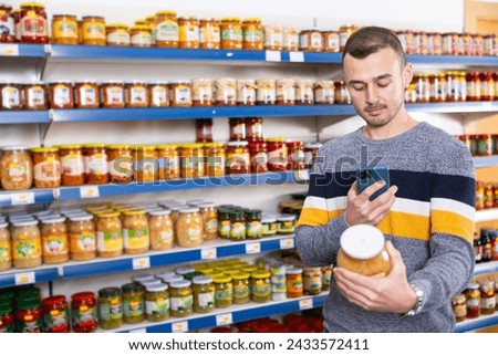 Man in store of imported Russian goods chooses jar with canned fermented cabbage. Male buyer photographs item, compares appearance of product in store and in mobile application