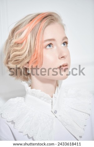 Beauty, style and fashion. A lovely teenage girl with a short haircut and blonde hair with orange streaks poses in a white blouse with a lace collar on a white background. 