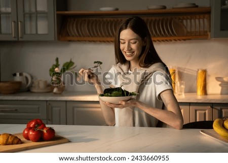 A smiling young woman is sitting at a kitchen counter bathed in morning light, about to enjoy a bowl of green salad. The kitchen is warmly lit and well-stocked, illustrating a healthy, domestic