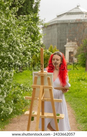 Female artist painting on plein air in botanical garden. Red long haired woman in lavender dress with wooden easel and canvas drawing outdoor. Glass greenhouse on background