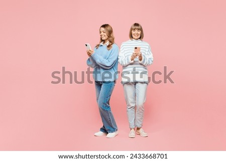 Full body smiling elder parent mom with young adult daughter two women together wear blue casual clothes hold use mobile cell phone isolated on plain pastel light pink background. Family day concept