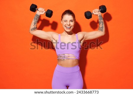 Young fitness trainer instructor woman sportsman wear purple top clothes spend time in home gym hold dumbbells raise up hands wink isolated on plain orange background. Workout sport fit abs concept