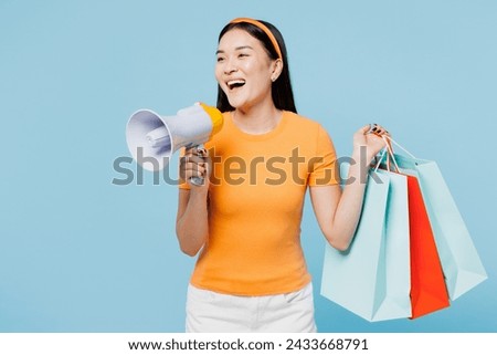 Young woman of Asian ethnicity wear orange t-shirt casual clothes hold in hand paper package bags after shopping scream in megaphone isolated on plain blue background Black Friday sale buy day concept