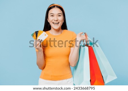 Young cool woman of Asian ethnicity wear orange t-shirt casual clothes hold package bags after shopping hold credit bank card isolated on plain blue cyan background. Black Friday sale buy day concept