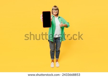 Elderly blonde woman 50s years old wears green shirt glasses casual clothes hold in hand use blank screen area mobile cell phone show thumb up isolated on plain yellow background. Lifestyle concept