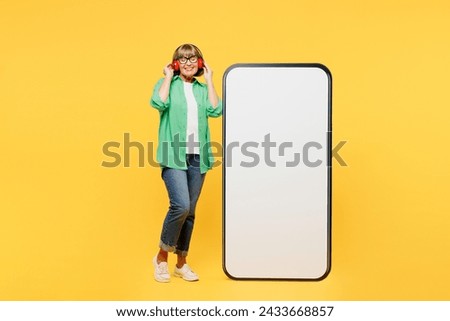 Full body elderly woman 50s year old wear green shirt glasses casual clothes big huge blank screen mobile cell phone smartphone with area listen music in headphones isolated on plain yellow background