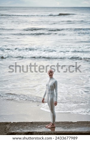 Young hairless girl with alopecia in white futuristic suit standing on concrete fence on sea background, bald pretty teenage girl symbolizes courage and acceptance of unique appearance