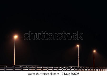 Street lampposts casts dim yellow warm along shore pier standing against pitch black canvas of night sky paints poetic picture of pier in stillness of night, serenity of solitary moments