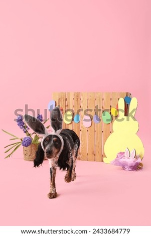 Cute cocker spaniel in bunny ears with Easter cake, paper rabbit, beautiful flowers and wooden fence on pink background